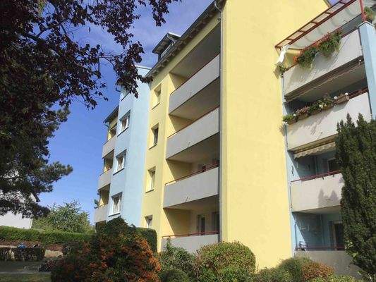 Rückansicht des mehrstöckigen Wohngebäudes mit Loggien (Balkonen), umgeben von Grünanlagen mit Bäumen und grünen Sträuchern