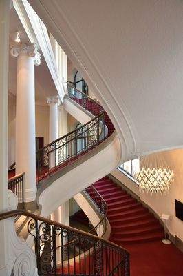 Elegantes Treppenhaus mit rotem Teppichläufer, weißen Säulen, geschwungenem Treppenverlauf und stilvoller Lichtinstallation.