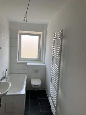 Badezimmer mit Badewanne, Handtuchheizkörper, Hänge-WC und Fenster. Fußboden anthrazit, Weiße Fliesen