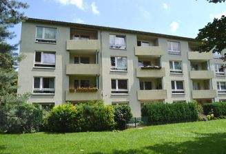 Rückansicht eines mehrstöckigen  Wohngebäudes mit Balkonen, umgeben von Bäumen und grünen  Sträuchern 