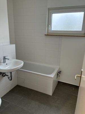 Badezimmer. Mit Fenster, neuen weißen Sanitärobjekten, weißen Wandfliesen und dunklen Bodenfliesen.