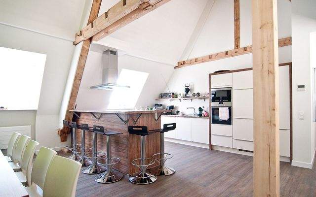 Moderne Büroküche mit Thekenbereich, fünf Barhockern, Kochfeld mit Dunstabzugshaube und sichtbarer Dachkonstruktion.