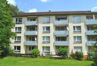 Rückansicht eines mehrstöckigen  Wohngebäudes mit Balkonen, umgeben von Bäumen und grünen  Sträuchern 