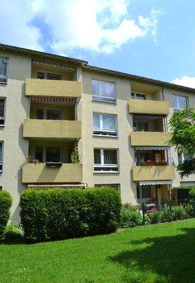 Rückansicht eines mehrstöckigen  Wohngebäudes mit Balkonen, umgeben von Bäumen und grünen  Sträuchern 