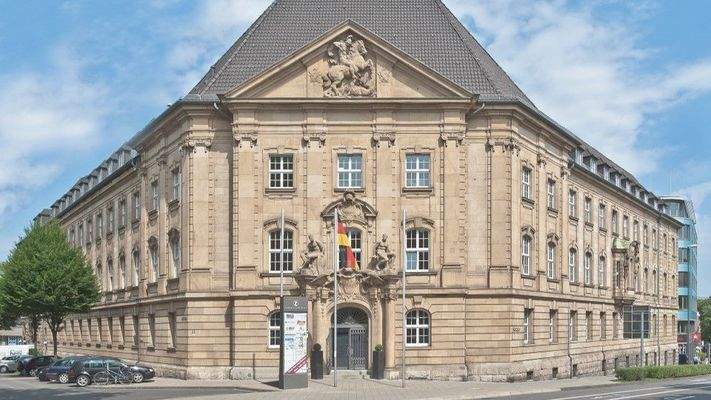 Außenansicht eines historischen Gebäudes mit Sandsteinfassade, aufwändiger Ornamentik und Walmdach, zentralem Eingangsportal und deutschen Flaggen.