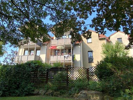 Rückansicht eines hellen mehrstöckigen Wohngebäudes mit Fenstern und Balkonen umgeben von einer grünen Außenanlage mit vielen Bäumen und Hecken