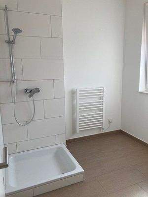 Tageslicht Bad mit Dusche mit weißem wandhängendem WC, Waschbecken und Handtuch Heizkörper. Die Wände sind im Nassbereich raumhoch und weiß gefliest. Der Boden ist hell gefliest.