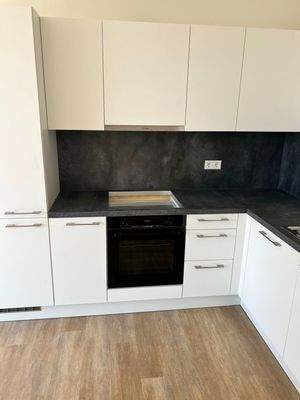 Beispiel einer L-förmigen Küche mit weißen Unterschränken und grifflose, helle Oberschränke. Die Arbeitsplatte sowie der Spritzschutz an der Wand sind aus einem anthrazitfarbenen Material. Der Boden ist ein Vinylboden in Holzoptik.