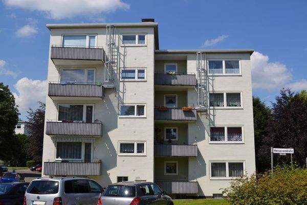 Rückansicht eines mehrstöckigen  Wohngebäudes mit Balkonen, umgeben von Bäumen und grünen  Sträuchern 