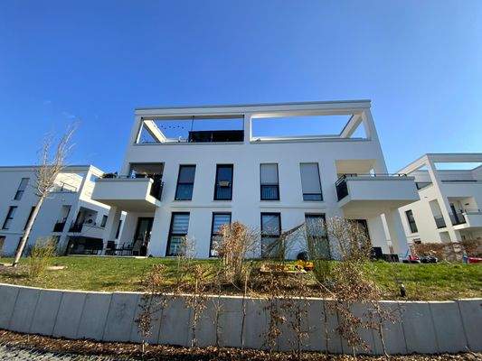 Rückansicht eines mehrstöckigen weißen Wohngebäudes mit Fenstern, Balkonen und Dachterrassen umgeben von einer grünen Außenanlage