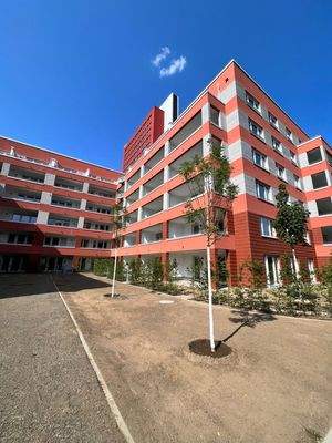 Rückansicht des mehrstöckigen Wohngebäudes mit Loggien und einer rot-grauen Fassadengestaltung. Innenhof mit jungen Bäumen und Sträuchern