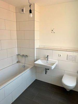 Beispiel eines Badezimmers mit weißem Wand-WC, weißem Wandwaschbecken und Badewanne mit weißen Wandfliesen auf anthrazitfarbenen Fliesenboden