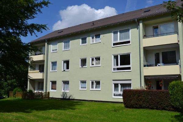  Rückansicht eines mehrstöckigen  Wohngebäudes mit Balkonen, umgeben von Bäumen und grünen  Sträuchern 