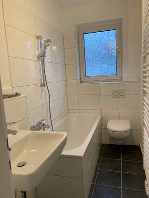 Tageslicht-Bad mit dunklen Bodenfliesen, weißen Wandfliesen, weißem Hänge-WC, weißem Waschbecken und einer Badewanne.