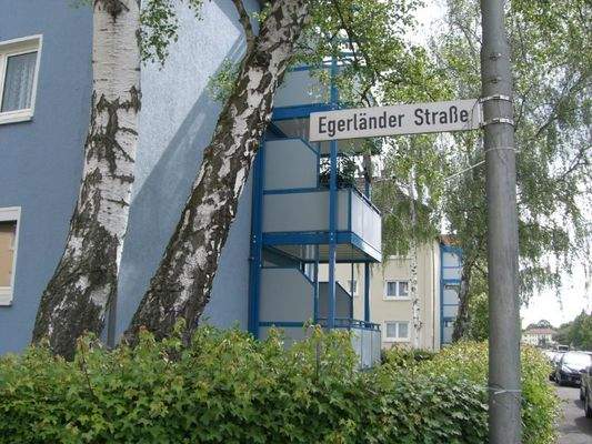 Seitenansicht eines mehrstöckigen blauben Gebäudes mit Balkonen, umgeben von einer grünen Hecke