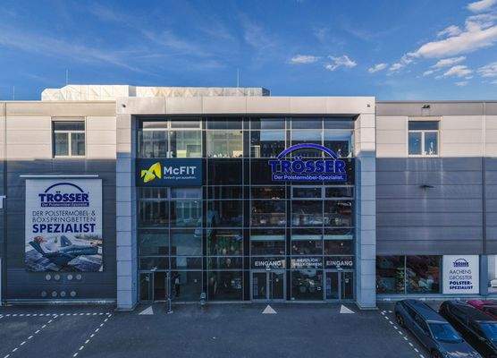 Frontalansicht eines modernen Gewerbegebäudes mit Glasfassade und den Logos von Trösser, dem Polstermöbel-Spezialisten, sowie McFIT Fitnessstudio. Zwei Eingänge mit Beschilderung ‚Herzlich willkommen‘ führen ins Gebäude. Im Vordergrund befinden sich Parkp