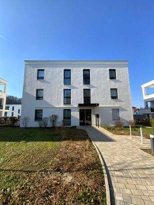 Vorderansicht eines mehrstöckigen weißen Wohngebäudes mit Fenstern, umgeben von einer grünen Außanlage mit Zuweg.