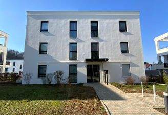 Vorderansicht eines mehrstöckigen weißen Wohngebäudes mit Fenstern, umgeben von einer grünen Außanlage mit Zuweg.