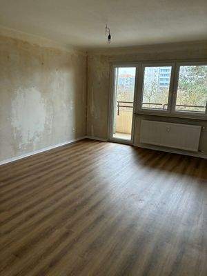 Blick in das Wohnzimmer. Mit Fenster und Zugang zum Balkon. Neuer brauner Vinylboden. Decken weiß angelegt. Wände tapezierfähig.