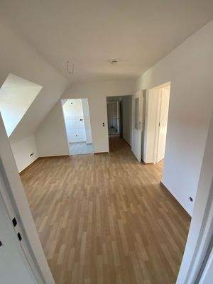 Wohnzimmer mit braunem PVC Boden, weißen Wänden, Dachschräge und Fenster mit Blick Richtung Küche und Flur.jpg