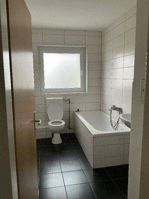 Badezimmer mit Badewanne und Fenster. Der Boden ist schwarz gefliest. Die Wände und die Sanitärobjekte sind weiß gehalten.