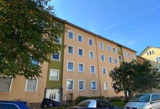 Voransicht eines mehrstöckigen hellorangenen Wohngebäudes mit Fenstern, umgeben von Bäumen und einer grünen Wiese an der Straße