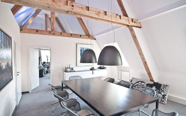 Besprechungsraum mit dunklem Holztisch, acht Designerstühlen, zwei großen schwarzen Pendelleuchten und offenen Holzbalken.