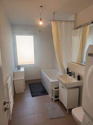 Tageslicht Wannen Badezimmer mit weißem wandhängenden WC, Waschbecken und Handtuchheizkörper. Die Wände sind im Nassbereich raumhoch und weiß gefliest. Der Boden ist hell gefliest.