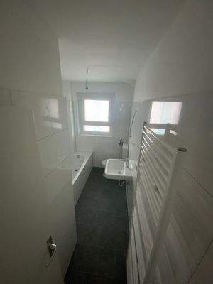 Modernisiertes Badezimmer mit Tageslicht, weißer Badewanne, weißem Wandwaschbecken, weißem Stand-WC, weißen Wandfliesen, Handtruchheizkörper und grauen Bodenfliesen