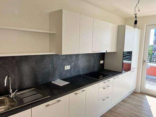 Beispiel einer Küche mit weißen Unterschränken und grifflose, helle Oberschränke. Arbeitsplatte und Spritzschutz sind in einem anthrazitfarbenen Material gehalten. Mittig ist ein Kochfeld eingebaut, rechts ein hoher Schrank mit integrieten Backofen.