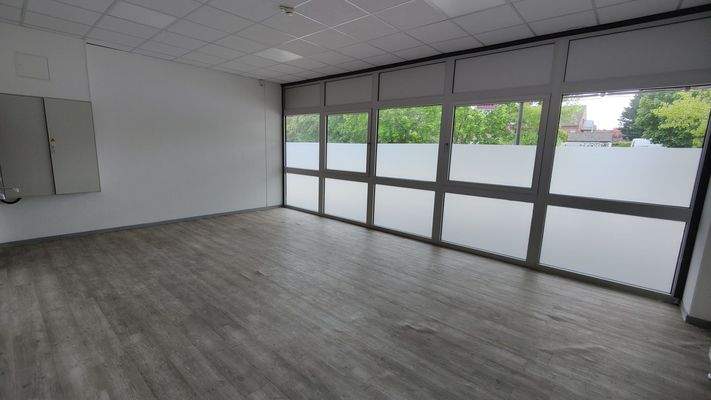 Heller Raum mit großflächiger Fensterfront, Milchglas im unteren Bereich, grauer Vinylboden und weißer Wand mit Elektroschrank.