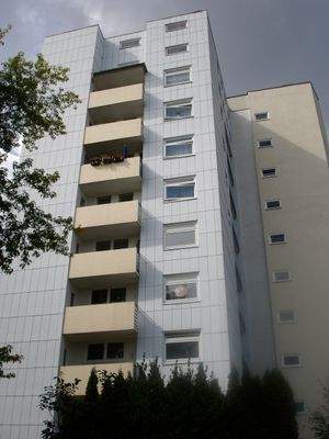 Rückansicht eines mehrstöckigen  Wohngebäudes mit Balkonen, umgeben von Bäumen und grünen  Sträuchern 