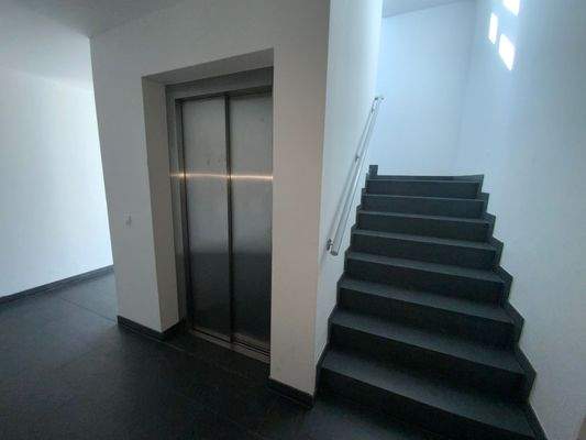 Blick auf silberne Aufzugstür und in Treppenhaus grauem Fliesenboden und weißen Wänden