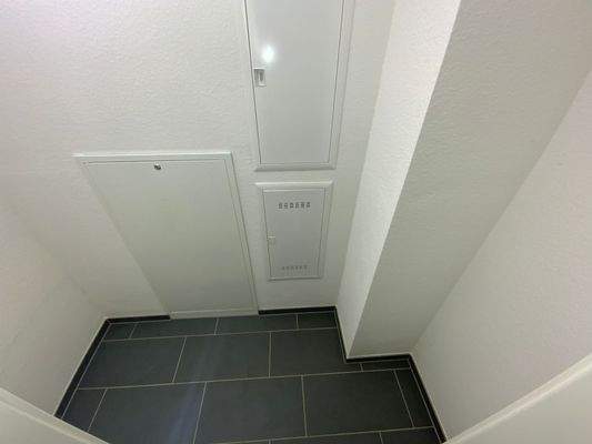 Abstellraum mit weißen Wänden und dunklen Bodenfliesen 