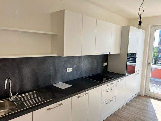 Beispiel einer Küche mit weißen Unterschränken und grifflose, helle Oberschränke. Arbeitsplatte und Spritzschutz sind in einem anthrazitfarbenen Material gehalten. Mittig ist ein Kochfeld eingebaut, rechts ein hoher Schrank mit integrieten Backofen.