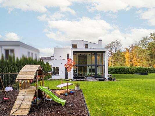 Modernes, weißes Haus mit großer Glasfront und Terrasse, davor ein Garten mit Spielplatz, Rasen und herbstlichen Bäumen im Hintergrund