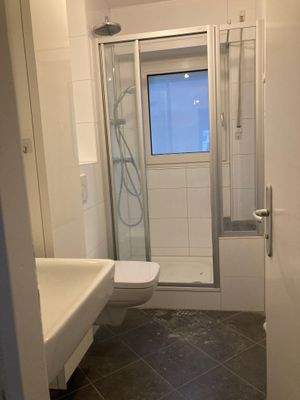 Tageslicht-Badezimmer mit anthrazitfarbenen Bodenfliesen und weißen Wandfliesen. Weißem Wand WC, Handwaschbecken und einer Dusche.