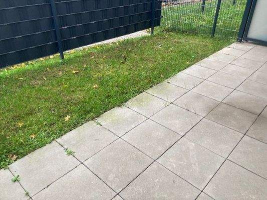 Blick von Balkontür in den Garten mit grauem Gartenzaun, helle Terrassenfliesen und Rasen