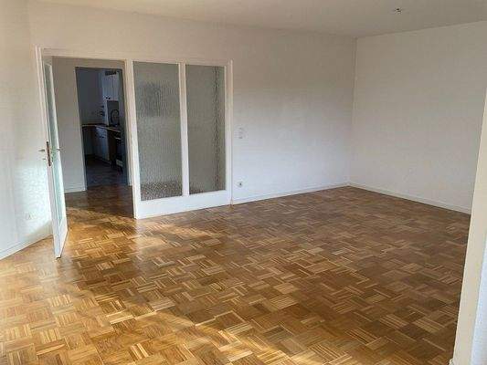 Wohnzimmer mit Parkettboden, weiße Wände und Zimmertür mit Glaselementen zum Flur