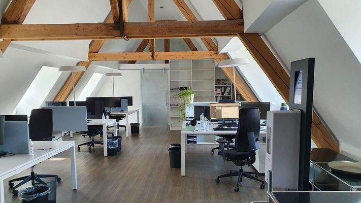 Großraumbüro unter dem Dach mit weißem Mobiliar, freiliegenden Holzträgern und modernen Bildschirmarbeitsplätzen.