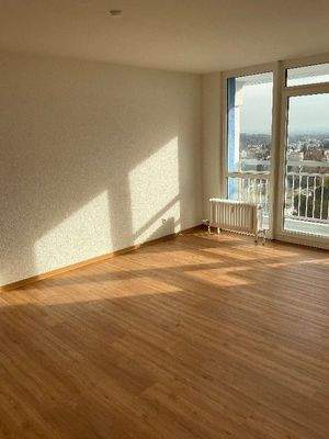 Ansicht des Wohzimmers mit weißen Wänden, Design-Vinylboden in Holzoptik, sowie Balkontür mit Zugang zum Balkon.