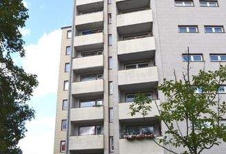 Rückansicht eines mehrstöckigen  Wohngebäudes mit Balkonen, umgeben von Bäumen und grünen  Sträuchern 