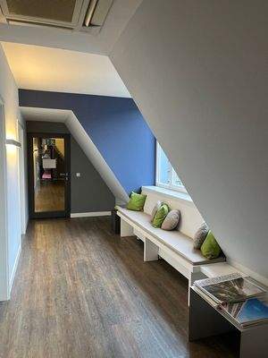 Flurbereich mit schräger Dachwand, Sitzbank mit Kissen unter dem Fenster, dunklem Holzfußboden und blau-grau abgesetzter Wandgestaltung.