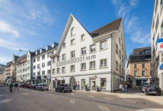 Rathausstraße Bregenz: Gebäude mit spitzem Giebel und der Aufschrift "0,0000000001 mm" auf der Fassade, Autos und Fußgänger auf der Straße.