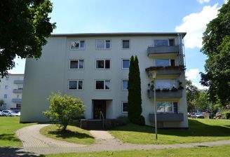 Vorderansicht eines mehrstöckigen  Wohngebäudes mit Balkonen, umgeben von Bäumen und grünen  Sträuchern 