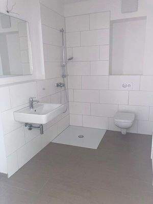 Tageslicht Dusch Badezimmer mit weißem wandhängendem WC, Waschbecken und Handtuch Heizkörper. Die Wände sind im Nassbereich Türhoch und weiß gefliest. Der Boden ist dunkel gefliest. 