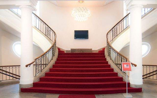 Zentraler Aufgang mit breiter roter Treppe, flankiert von weißen Säulen und geschwungenem Geländer, beleuchtet durch modernen Kronleuchter.