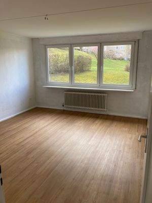 Wohnzimmer mit weiß grundierten Wänden, PVC Boden in Holzoptik und großen Fenstern mit Blick in die grüne Außenanlage