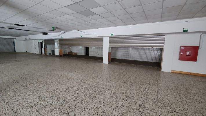 Großer Raum mit Deckenplatten, Rollgittern und gekachelter Rückwand, geeignet als Lager- oder Verkaufsfläche.