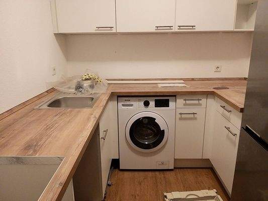 Küche, hellbraune Arbeitsplatte, weiße Küchenschränke, Herd, Spülbecken, Kühlschrank und Boden in Holzoptik